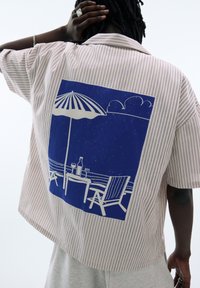 Camicia a maniche corte con righe verticali, caratterizzata da una grafica blu di una scena da spiaggia con sedie, tavolo, bottiglia e ombrellone sul retro.