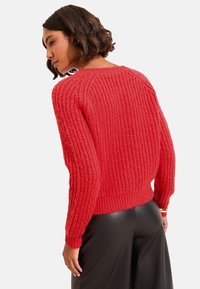 Maglione rosso lavorato a maglia con un motivo testurizzato, polsini e orlo a coste, abbinato a pantaloni neri a vita alta, che esprimono una vestibilità comoda.