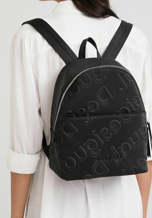 Woman in white shirt wearing black backpack with raised repeating logo pattern and silver zipper, shown from the back.