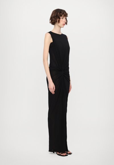 Vestido maxi negro sin mangas con un detalle de nudo torcido en la cintura, textura suave y silueta ajustada, combinado con sandalias negras con tiras.