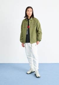 Olijfgroene lichtgewicht jas met zwart logo, gecombineerd met een zwart t-shirt, witte joggers en beige sneakers. Casual streetwear stijl.