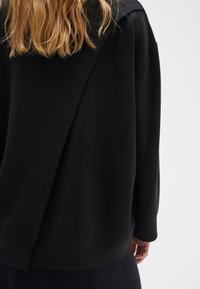 Sudadera negra de gran tamaño con un ajuste relajado, textura suave, hombros caídos y un diseño distintivo en la parte posterior con un detalle drapeado.