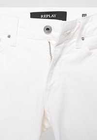 Jean en coton blanc avec fermeture éclair et bouton en métal. Étiquette "REPLAY" à la ceinture, avec des détails de couture sur les poches et les coutures.