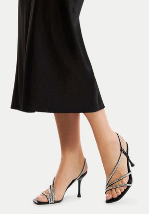 Stopy w czarnych sandałach na wysokim obcasie z paskami ozdobionymi kryształkami, zestawione z czarną spódnicą midi na jednolitym jasnym tle.