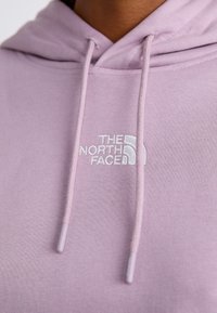 Gros plan sur une personne portant un hoodie violet clair avec le logo brodé blanc "The North Face" et des cordons assortis.