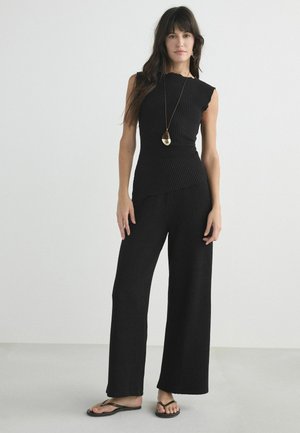 Frau steht, trägt ein ärmelloses schwarzes geripptes Oberteil und weite Hosen mit schwarzen Flip-Flops und eine lange Halskette mit Anhänger.