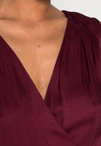 Detailní záběr na horní část hrudníku a krku osoby s viditelnou vertikální jizvou, oblečenou v tmavě burgundovém výstřihu do V.