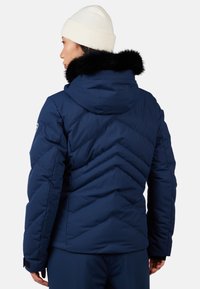 Giacca puffer blu navy con design a zigzag, dotata di un colletto in pelliccia nera removibile e polsini regolabili. Indossata con un berretto lavorato a maglia bianco.