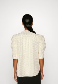 Femme aux cheveux foncés attachés en queue basse, portant un chemisier blanc à paillettes dorées et manches bouffantes, debout devant un fond uni.