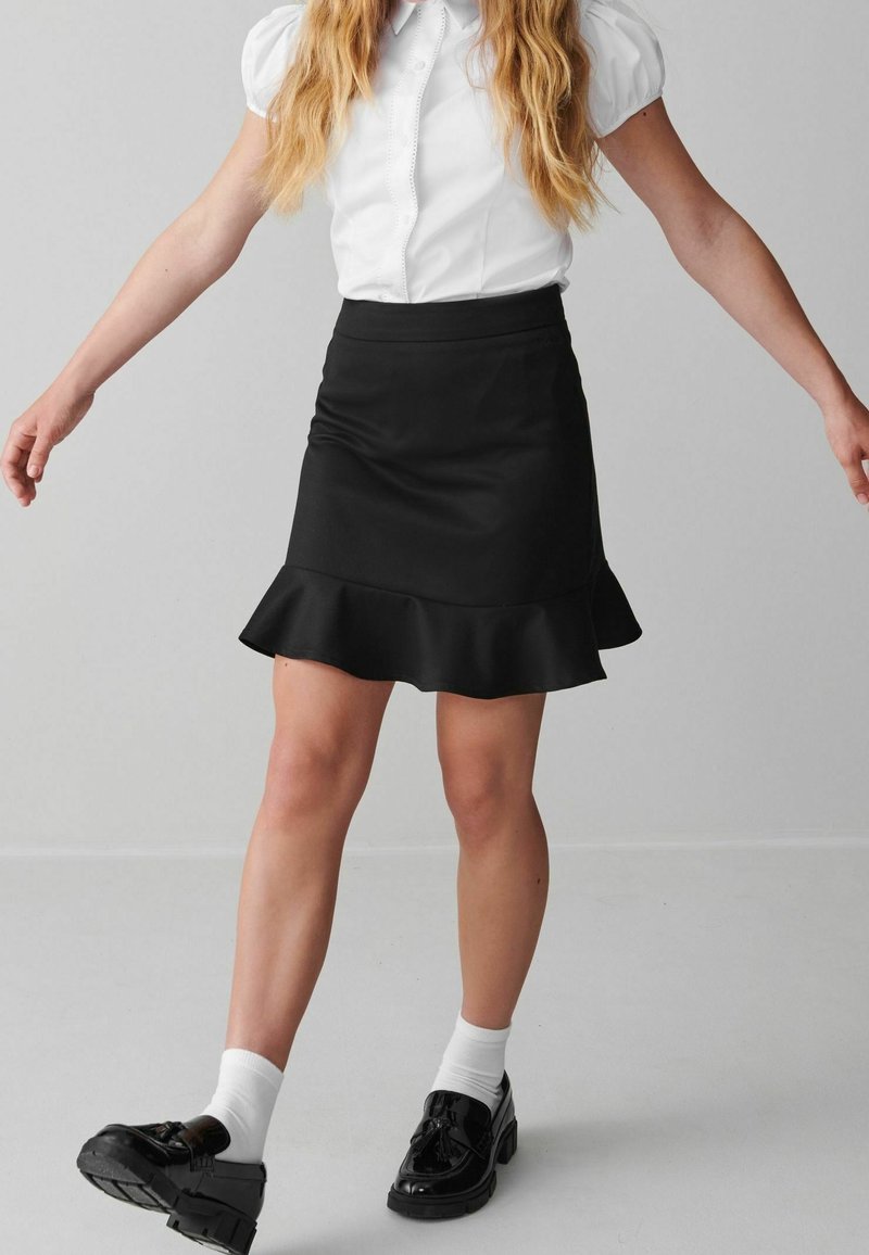 Femme portant un chemisier blanc à manches bouffantes, une jupe noire avec un ourlet à volants, des chaussettes blanches et des mocassins noirs, debout devant un fond gris uni.