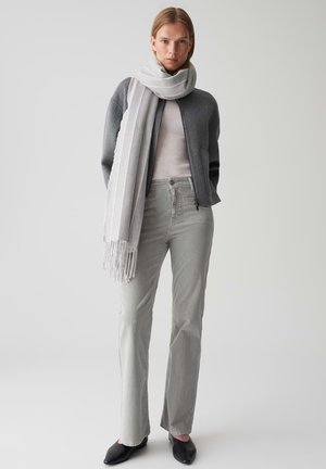 Cardigan en laine gris, haut gris clair et jean gris clair. Chaussures plates noires à bout pointu. Écharpe rayée blanche avec des détails à franges. Textures lisses.