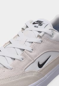 Κοντινό πλάνο λευκού και μπεζ αθλητικού παπουτσιού Nike SB με λευκά κορδόνια, μαύρο λογότυπο swoosh στο πλάι και κείμενο "Nike SB Valor" στην ετικέτα της γλώσσας.