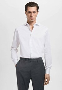 Camisa de vestir blanca de mangas largas con cuello, combinada con pantalones oscuros de corte entallado en gris. La tela es suave, con detalles de botones en la parte delantera.