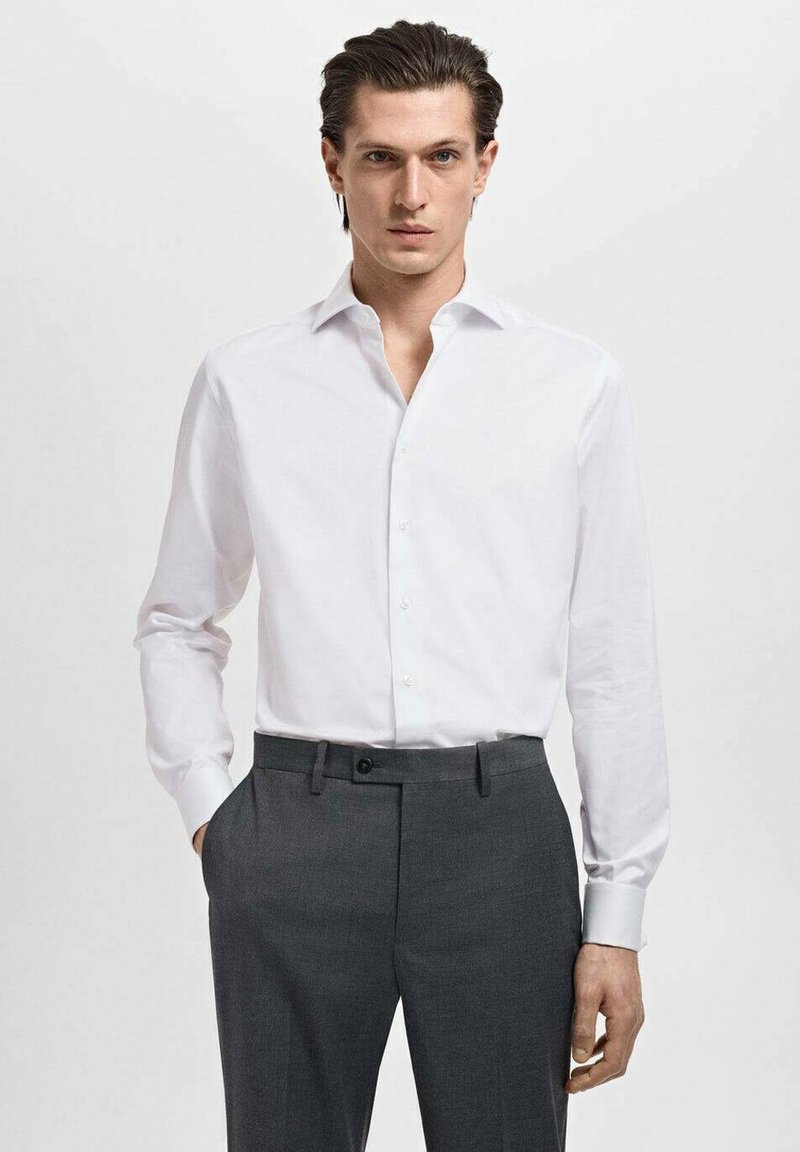 Camisa de vestir blanca de mangas largas con cuello, combinada con pantalones oscuros de corte entallado en gris. La tela es suave, con detalles de botones en la parte delantera.