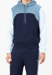 Navyblå hoodie med ljusblå axlar, känguruficka och dragsnören. Har Lacoste-logotyp på bröstet, material i bomull.