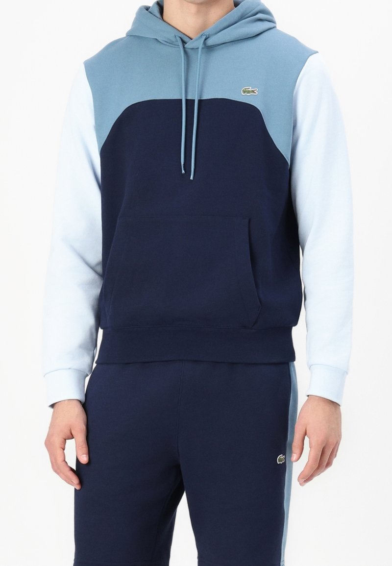 Navyblå hoodie med ljusblå axlar, känguruficka och dragsnören. Har Lacoste-logotyp på bröstet, material i bomull.
