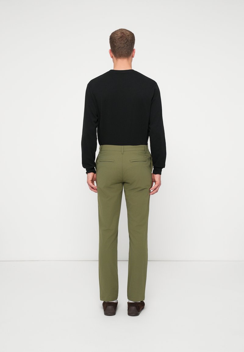 Maglietta nera a maniche lunghe con silhouette aderente, abbinata a pantaloni tailored verde oliva con tasche posteriori e una texture liscia. Scarpe marroni.