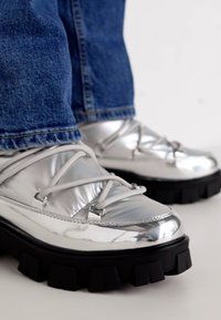Sølvreflekterende ankelstøvler med gummisåle, med snøring og polstret design. Brukt med mørke denimjeans.