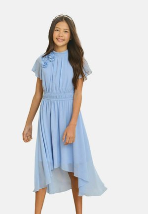 Meisje met lang donker haar dat een lichtblauwe high-low jurk met bloemenpatroon en een parelhaarband draagt, glimlachend naar de camera.