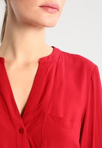 Blouse rouge à manches courtes avec un col en V, une fermeture à un bouton et une poche poitrine gauche. Tissu lisse avec une finition légèrement brillante.