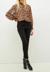 Zwarte hoge taille broek gecombineerd met een doorschijnende blouse met bloemmotief, lange mouwen en een hoge halslijn. Glanzende zwarte enkellaarsjes.