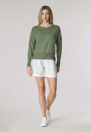 Maglione a maglia verde oliva con scollatura rotonda, polsini e orlo a coste, abbinato a pantaloni corti in lino bianchi. Indossato con sneakers bianche.