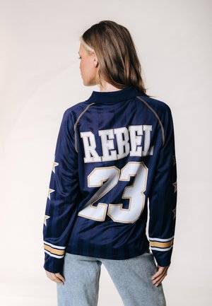 Femme de dos portant un maillot bleu marine avec des étoiles blanches et dorées et "REBEL 23" dans le dos, associée à un jean bleu clair.