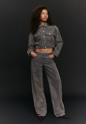Young woman with curly hair wearing a textured gray cropped jacket and matching wide-leg pants, standing with hands in pockets against dark background.