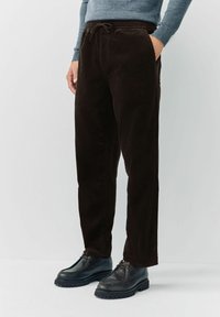 Man wearing dark brown corduroy pants with drawstring waist and black leather shoes, standing against a plain light background.