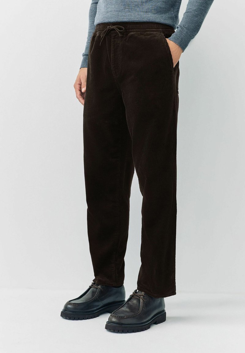 Man wearing dark brown corduroy pants with drawstring waist and black leather shoes, standing against a plain light background.