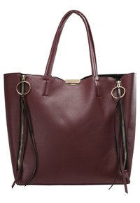 Grand sac cabas en cuir rouge foncé avec double poignée, fermetures éclair latérales verticales avec anneaux ronds et plaque de marque dorée en haut au centre.