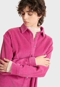 Camisa de pana fucsia con diseño de botones, mangas largas y textura acanalada; cuenta con un cuello extendido y botones a juego.