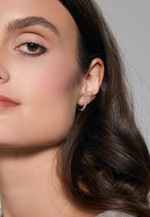 Srebrna kolczyk w kształcie obręczy z przezroczystymi, błyszczącymi akcentami; gładka powierzchnia; noszony na małżowinie usznej, w połączeniu z falującymi, ciemno-brązowymi włosami i neutralnym odcieniem skóry.