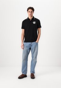 Schwarzes Poloshirt mit weißen Kragenakzenten und gesticktem Logo, kombiniert mit hellblauen Jeans und braunen Schuhen, vor einem weißen Hintergrund.