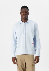 Blue and white checkered button-up shirt with long sleeves, featuring a visible red logo on the left side and a softened collar design.