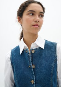 Donkerblauw denim vest met een gestructureerd ontwerp, voorzien van een knoopsluiting, zichtbare stiksels, en een witte overhemd met kraag eronder.