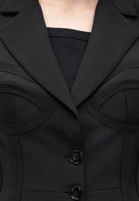 Κοντινό πλάνο μαύρου προσαρμοσμένου σακακιού με δομημένες ραφές πάνω από το στήθος και δύο μαύρα κουμπιά κλεισμένα στο μπροστινό μέρος.