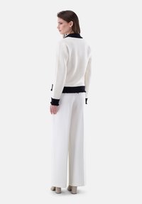 Weißer strukturierter Cardigan mit schwarzem Besatz und fransigen Kanten, kombiniert mit weißen weiten Hosen und beigen Blockabsatzschuhen.