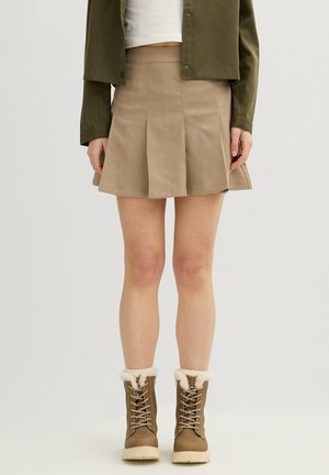 Persona con minifalda plisada beige, chaqueta verde oliva, top corto blanco y botas marrón con cordones y forro de borrego, de pie sobre un fondo liso.