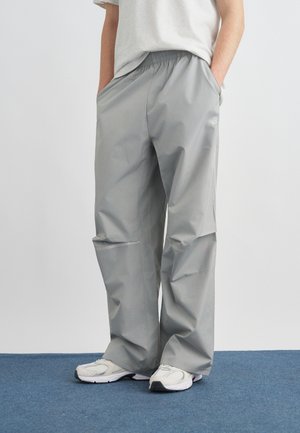 Oseba, oblečena v ohlapne sive hlače z elastičnim pasom, bele superge in belo majico, z rokami v žepih, stoji na modrem preprogi.