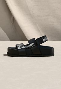 Μαύρο σανδάλι πλατφόρμα με το λογότυπο Moschino στα λουράκια, με δύο μπροστινές λωρίδες και μια ρυθμιζόμενη πίσω λουρίδα σε ουδέτερη επιφάνεια.