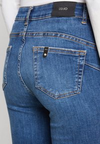 Vista trasera de unos pantalones vaqueros de mezclilla azul con costuras naranjas, bolsillos traseros, una etiqueta de marca negra con la inscripción "LIU•JO" y una pequeña etiqueta de metal de la marca.