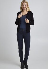 Mustat neuletakki, jossa on pyöreä pääntie ja nappikiinnitys, yllä tummansininen paita ja tummat skinny-farkut, yhdistettynä nilkkureihin.