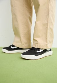 Svart sneakers med hvite detaljer, med en glatt syntetisk overdel, rund form og gummisåle, kombinert med lyse beige bukser.