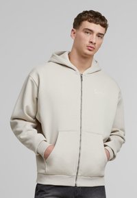 Hettegenser med glidelås i lys beige bomullsblanding, med frontlomme, ribbestrikkede mansjetter og en liten hvit logo på brystet.