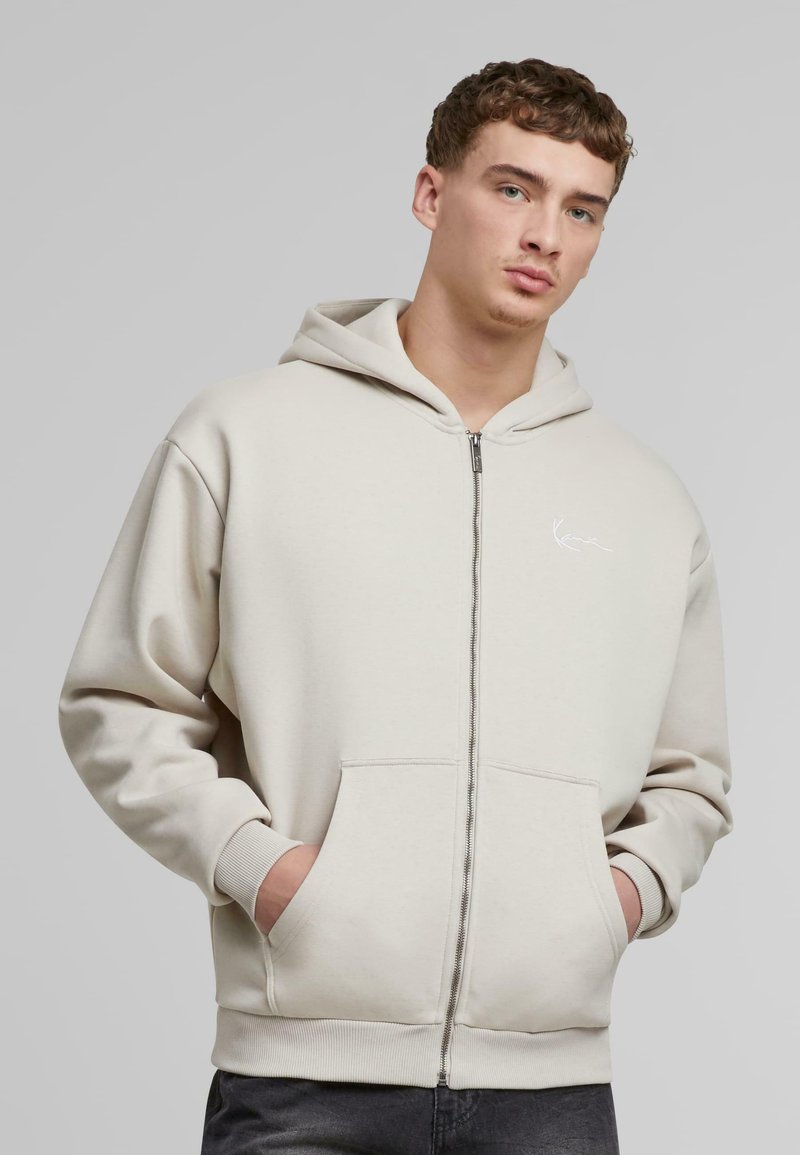 Hettegenser med glidelås i lys beige bomullsblanding, med frontlomme, ribbestrikkede mansjetter og en liten hvit logo på brystet.