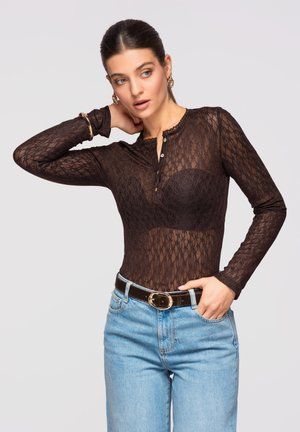 Vrouw draagt een zwarte doorschijnende kanten top met lange mouwen en knopen, zwarte bh eronder, lichtblauwe spijkerbroek, zwarte riem, gouden oorringen en armband.