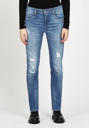 Person in hellblauen, distressed Skinny Jeans, schwarzem Langarmoberteil und schwarzen Schnürstiefeln vor einem schlichten Hintergrund.