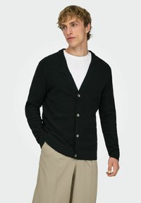 Man with blond hair wearing a black buttoned cardigan over a white shirt and beige pants, looking slightly to the side with one hand in pocket.