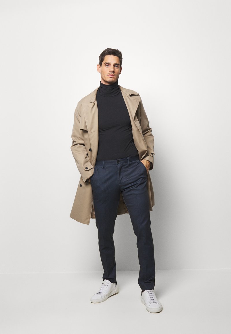 Abrigo trench de color beige, cuello alto negro, pantalones azul marino texturizados y zapatillas blancas. El abrigo cuenta con botones y un corte holgado.